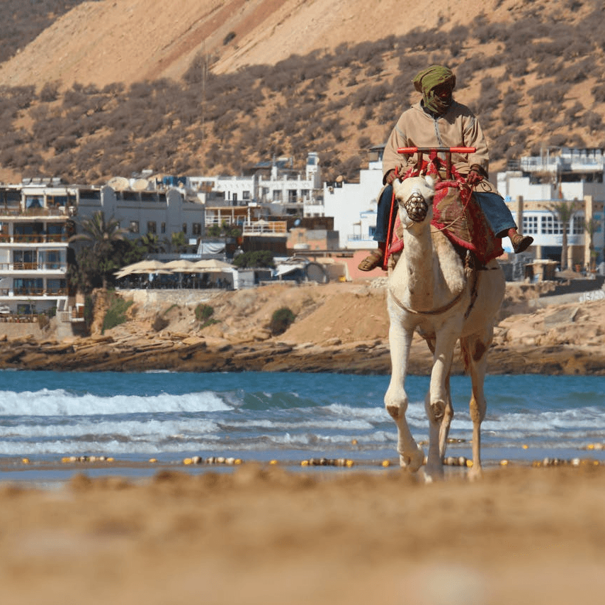 A beautiful coastal view of Taghazout, Morocco, showing surfers riding the waves and golden sandy beaches, perfect for travelers exploring the area.